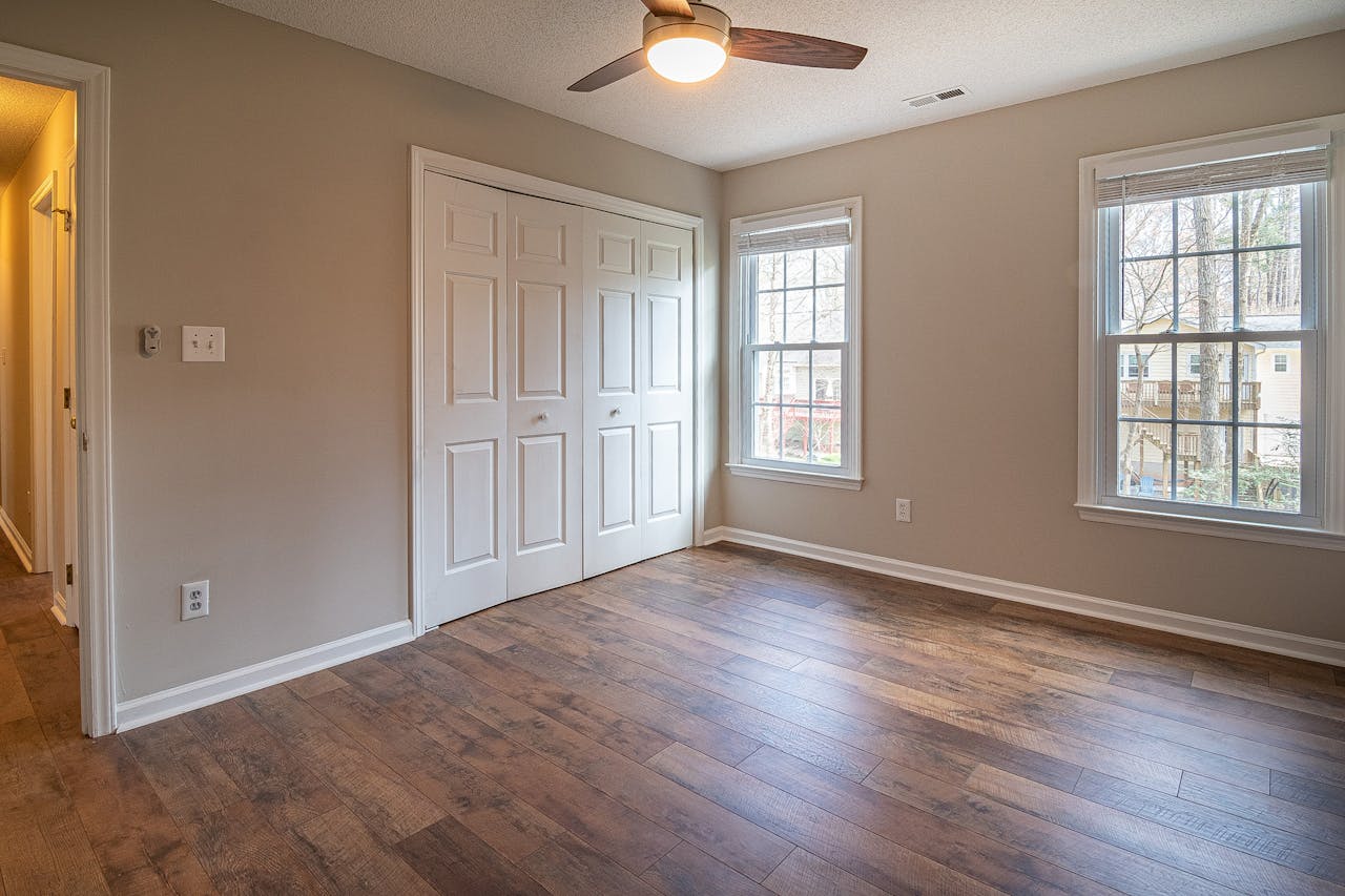 gallery-1 Spacious empty room with hardwood floors, ceiling fan, and natural light from windows.