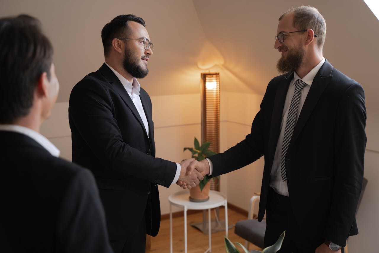 gallery-5 Two businessmen in suits shaking hands in a warm office setting.